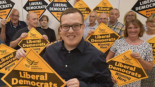 Jack Davies holding a Lib Dem diamond poster saying "winning here" with a group of members behind